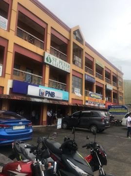 A commercial building with multiple signs indicating various businesses, such as 'St. Peter Life Plan' and 'PNB'. Cars and motorcycles are parked in front. The structure is three stories high, with a yellow and brown facade. Pedestrians are walking on the sidewalk.