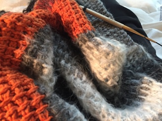 A close-up view of textured knitting work in various colors including orange, gray, and cream. A single knitting needle is partially inserted into the fabric, suggesting ongoing work. The fibers appear soft and woolly.