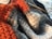 A close-up view of textured knitting work in various colors including orange, gray, and cream. A single knitting needle is partially inserted into the fabric, suggesting ongoing work. The fibers appear soft and woolly.
