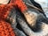 A close-up view of textured knitting work in various colors including orange, gray, and cream. A single knitting needle is partially inserted into the fabric, suggesting ongoing work. The fibers appear soft and woolly.