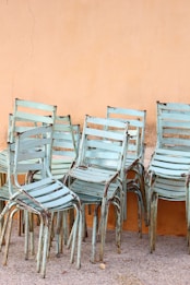 Several stacks of vintage metal chairs with a distressed turquoise paint finish, arranged against a cracked, peach-colored wall on a concrete floor.