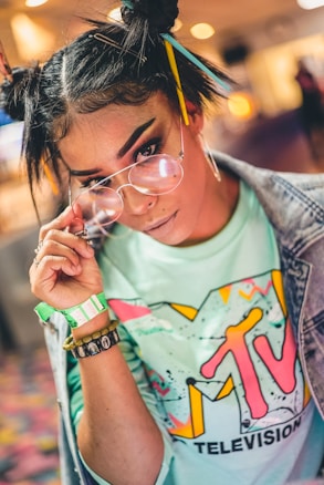 A person with an edgy hairstyle featuring chopsticks and bobby pins is wearing large round glasses, a mint green shirt with a colorful vintage MTV logo, and a denim jacket. The background is softly blurred with warm lighting.