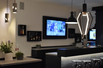 A modern bar interior with contemporary lighting fixtures and a sleek counter design. Wall-mounted shelves hold bottles, and a large screen displays a blurred image. Potted plants add a touch of greenery, and bar stools line the counter.