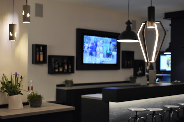 A modern bar interior with contemporary lighting fixtures and a sleek counter design. Wall-mounted shelves hold bottles, and a large screen displays a blurred image. Potted plants add a touch of greenery, and bar stools line the counter.