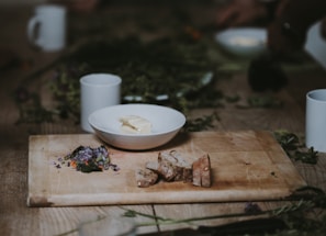 A rustic wooden table with a cutting board featuring sliced bread and a small pile of edible flowers. Beside it sits a white bowl containing a slab of butter. The scene is surrounded by scattered herbs and cups, creating a cozy and natural atmosphere.