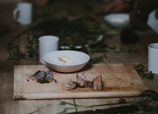 A rustic wooden table with a cutting board featuring sliced bread and a small pile of edible flowers. Beside it sits a white bowl containing a slab of butter. The scene is surrounded by scattered herbs and cups, creating a cozy and natural atmosphere.