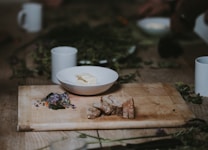 A rustic wooden table with a cutting board featuring sliced bread and a small pile of edible flowers. Beside it sits a white bowl containing a slab of butter. The scene is surrounded by scattered herbs and cups, creating a cozy and natural atmosphere.