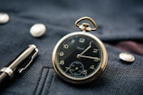 A vintage pocket watch with a black face and gold casing rests on a textured dark blue fabric, accompanied by a fountain pen nearby. The watch shows Roman numerals and intricate designs, with shiny metallic buttons on the fabric.