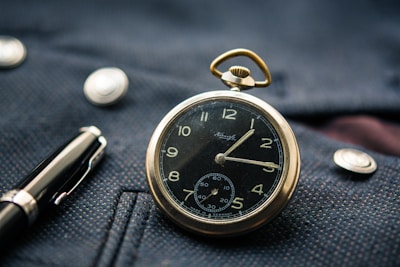 A vintage pocket watch with a black face and gold casing rests on a textured dark blue fabric, accompanied by a fountain pen nearby. The watch shows Roman numerals and intricate designs, with shiny metallic buttons on the fabric.