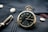 A vintage pocket watch with a black face and gold casing rests on a textured dark blue fabric, accompanied by a fountain pen nearby. The watch shows Roman numerals and intricate designs, with shiny metallic buttons on the fabric.