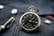 A vintage pocket watch with a black face and gold casing rests on a textured dark blue fabric, accompanied by a fountain pen nearby. The watch shows Roman numerals and intricate designs, with shiny metallic buttons on the fabric.