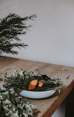 A rustic wooden table holds a white bowl filled with a variety of dried herbs mixed with a few vibrant orange flowers. The background is neutral, providing a simple and natural setting. Some green leafy branches extend from the left side.