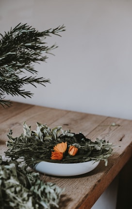 A rustic wooden table holds a white bowl filled with a variety of dried herbs mixed with a few vibrant orange flowers. The background is neutral, providing a simple and natural setting. Some green leafy branches extend from the left side.