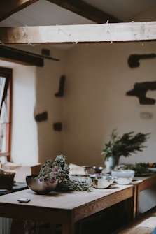 A rustic kitchen interior featuring a wooden table cluttered with metal bowls and bundles of dried herbs. Soft natural light filters through a window, casting gentle shadows on the table. In the background, there are decorative elements on a light-colored wall and sparse wooden beams under the ceiling.