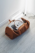 A brown leather duffel bag is placed on a light wood floor, partially open with a laptop, glasses, and a charger visible inside. Nearby, there are two standing fluorescent tube lights and visible cords on the floor.
