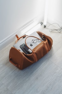 A brown leather duffel bag is placed on a light wood floor, partially open with a laptop, glasses, and a charger visible inside. Nearby, there are two standing fluorescent tube lights and visible cords on the floor.