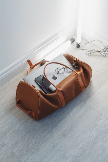 A brown leather duffel bag is placed on a light wood floor, partially open with a laptop, glasses, and a charger visible inside. Nearby, there are two standing fluorescent tube lights and visible cords on the floor.