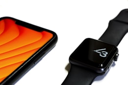 A close-up view of a smartphone and a smartwatch placed on a white surface. The smartphone has a bright orange screen, while the smartwatch displays the time with black and white color tones. Both devices have a sleek, modern design with black exterior finishes.