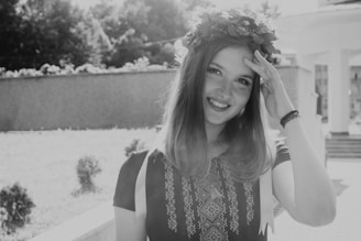 A young woman with long hair smiles warmly at the camera. She wears a floral wreath on her head and a dark, embroidered dress. Her hand is gently resting on her head. The background is outdoors with a bright, sunny setting, featuring greenery and a building structure.