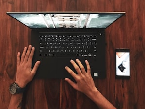 A person analyzing a smartphone and laptop side by side on a wooden desk.