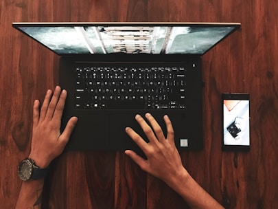 A person analyzing a smartphone and laptop side by side on a wooden desk.