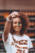 A person with curly hair is smiling and holding up a geometric pendant in focus. The background is blurred, emphasizing the jewelry piece.
