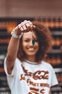 A person with curly hair is smiling and holding up a geometric pendant in focus. The background is blurred, emphasizing the jewelry piece.