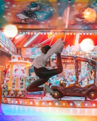 A young person is captured in mid-air performing a dance move in front of a colorful, illuminated amusement park ride. The background features vibrant lights and cartoon characters, creating a lively and playful atmosphere.
