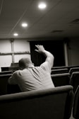 A person sitting in a church pew with their arm raised, facing the altar where a wooden cross is displayed. The setting is dimly lit with soft overhead lights and appears to be a place of worship.