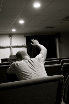 A person sitting in a church pew with their arm raised, facing the altar where a wooden cross is displayed. The setting is dimly lit with soft overhead lights and appears to be a place of worship.