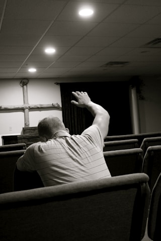 A person sitting in a church pew with their arm raised, facing the altar where a wooden cross is displayed. The setting is dimly lit with soft overhead lights and appears to be a place of worship.