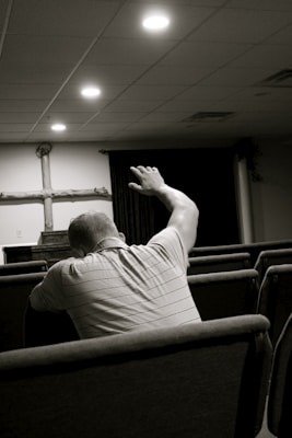 A person sitting in a church pew with their arm raised, facing the altar where a wooden cross is displayed. The setting is dimly lit with soft overhead lights and appears to be a place of worship.