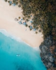 Aerial view of a pristine beach with turquoise water gently meeting the sandy shore. Palm trees cast shadows on the beach, where several thatched-roof huts and colorful umbrellas are visible. A lone boat floats on the clear blue sea, adding a sense of tranquility to the tropical scenery.