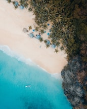 Aerial view of a pristine beach with turquoise water gently meeting the sandy shore. Palm trees cast shadows on the beach, where several thatched-roof huts and colorful umbrellas are visible. A lone boat floats on the clear blue sea, adding a sense of tranquility to the tropical scenery.