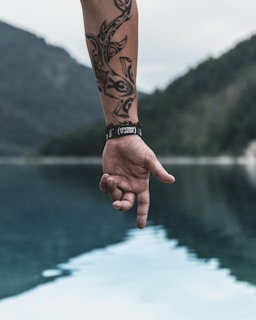 A tattooed arm extends over a body of water, with a wristband featuring a motivational phrase. The serene background includes blurred mountains and a reflective lake.
