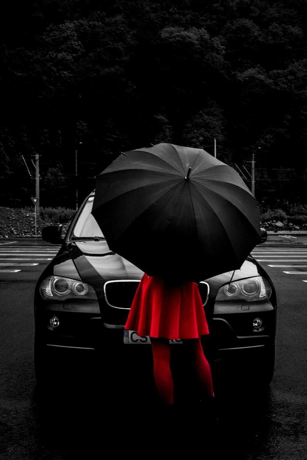 검은 우산 아래 빨간 플레어 드레스를 입은 여자가 차량 앞에 서 있습니다.