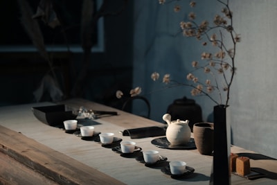 A rustic and minimalistic tea setup on a wooden table includes several small white ceramic cups on dark saucers, a white teapot, and a brown cup. A sprig with dried flowers stands in a simple black vase. There's a serene and calm ambiance with soft natural lighting casting shadows.