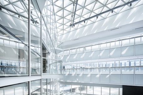 A modern and spacious architectural interior features extensive use of glass and steel. The structure has multiple levels with large glass panels that allow natural light to pour in, creating patterns of light and shadows. The ceiling design includes a geometric pattern that enhances the contemporary aesthetic.