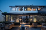 A modern two-story house with large windows and a tiled roof, surrounded by a well-maintained garden with bamboo and modern wicker patio furniture. The house is warmly lit, contrasting with the blue hues of the evening sky.