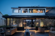 A modern two-story house with large windows and a tiled roof, surrounded by a well-maintained garden with bamboo and modern wicker patio furniture. The house is warmly lit, contrasting with the blue hues of the evening sky.