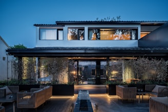 A modern two-story house with large windows and a tiled roof, surrounded by a well-maintained garden with bamboo and modern wicker patio furniture. The house is warmly lit, contrasting with the blue hues of the evening sky.