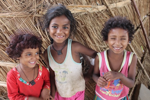 Three smiling children with dark hair stand in front of a wall made from straw or reeds. The child on the left wears a red shirt, the middle child wears a white tank top, and the child on the right wears a pink shirt. Their expressions convey joy and happiness.