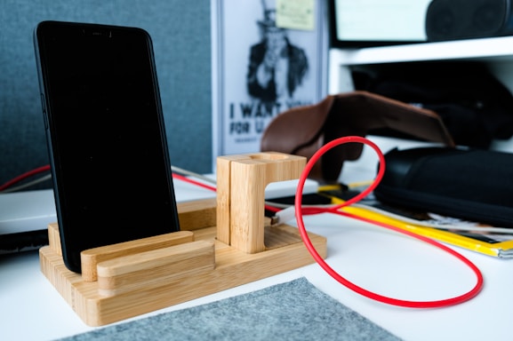 A smartphone is placed on a wooden stand with a red cable attached. In the background, there's a brown leather pouch and some documents. The setting appears to be an office desk with various items and a soft gray felt mat.