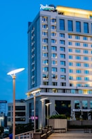 A tall, modern hotel building with large windows and illuminated signs stands against a clear evening sky. Streetlights in the foreground add a warm glow to the urban scene.