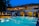 A luxurious hotel with a large outdoor swimming pool illuminated in blue light. The building has two stories with expansive windows revealing a warmly lit and elegant interior. There are decorative elements on the roof and a small bar area beside the pool. Palm trees are visible on the side, enhancing the tropical ambiance.