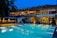 A luxurious hotel with a large outdoor swimming pool illuminated in blue light. The building has two stories with expansive windows revealing a warmly lit and elegant interior. There are decorative elements on the roof and a small bar area beside the pool. Palm trees are visible on the side, enhancing the tropical ambiance.