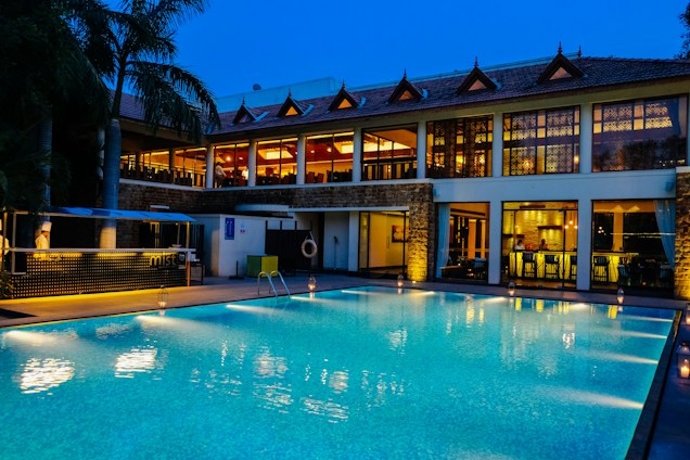 A luxurious hotel with a large outdoor swimming pool illuminated in blue light. The building has two stories with expansive windows revealing a warmly lit and elegant interior. There are decorative elements on the roof and a small bar area beside the pool. Palm trees are visible on the side, enhancing the tropical ambiance.
