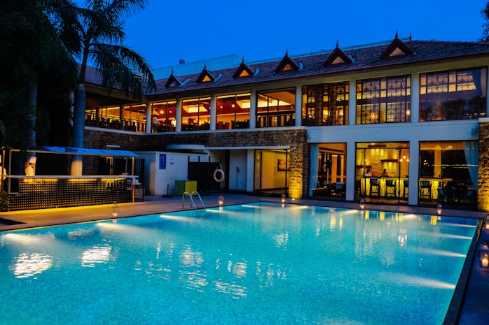 A luxurious hotel with a large outdoor swimming pool illuminated in blue light. The building has two stories with expansive windows revealing a warmly lit and elegant interior. There are decorative elements on the roof and a small bar area beside the pool. Palm trees are visible on the side, enhancing the tropical ambiance.