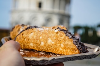 A hand is holding a dish with a cannoli pastry. The pastry has a golden-brown crispy shell with chocolate sprinkles on one end and powdered sugar dusted on top. The background features a blurred architectural structure, suggesting an outdoor setting.