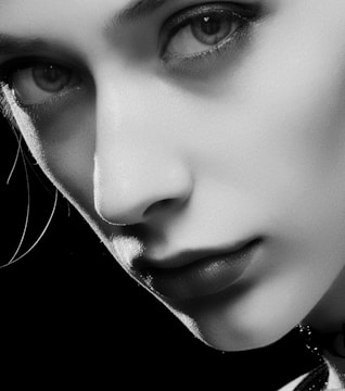 A black and white close-up portrait of a person's face, focusing on the eyes and lips. The smooth skin texture and subtle shadows emphasize facial features, creating a dramatic and intimate mood.